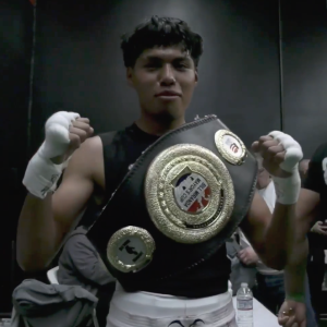 Gabriel Velasco, a fighter who competed at the event poses wearing white shorts with a black shirt with his title belt with a black strap nd gold plate.